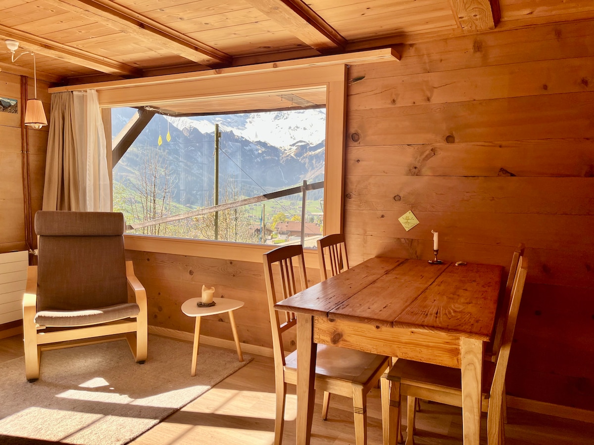 A cozy corner of the living room features a wooden table accompanied by three chairs. A comfortable armchair is positioned nearby. Natural light floods through a large panoramic window, offering a breathtaking view of the mountains, while warm wooden walls create an inviting atmosphere.