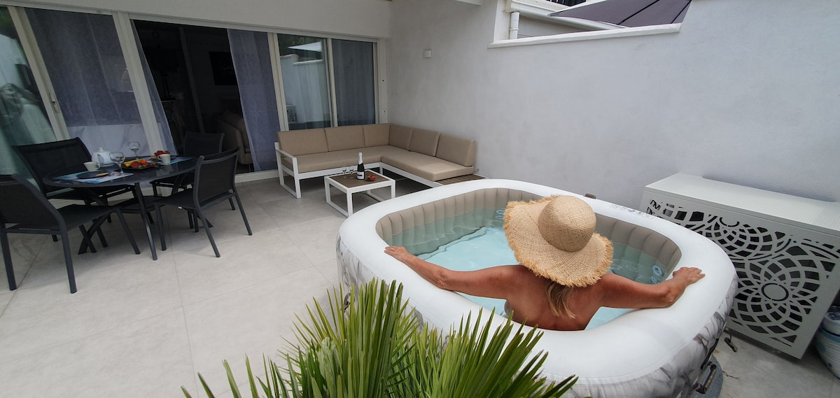 An outdoor terrace features a portable hot tub, with a person relaxing inside. Comfortable seating is arranged nearby, alongside a dining table set for two. The area is surrounded by potted greenery, and large sliding doors lead to the interior space.