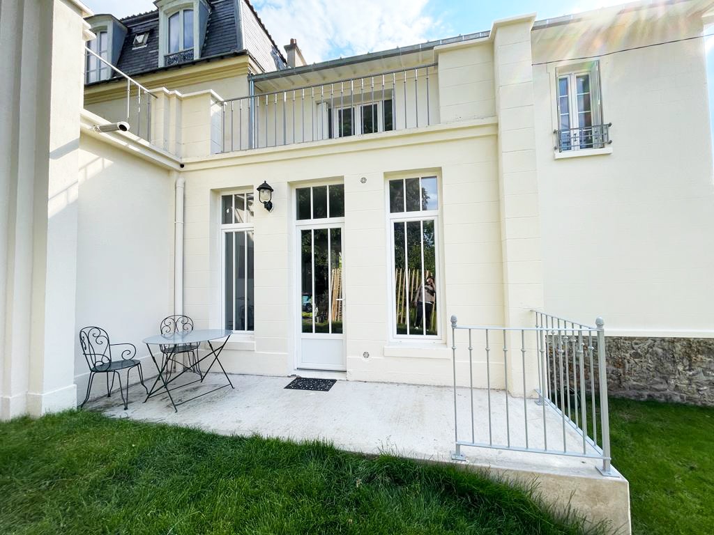 The image shows a charming outdoor terrace with a small metal table and two matching chairs. The patio is surrounded by manicured grass and features large windows that allow natural light to fill the interior. A decorative railing adds a touch of elegance.