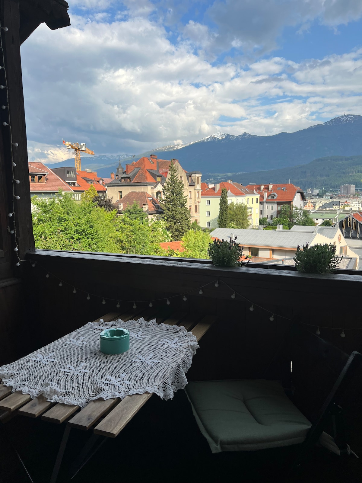 A small balcony is adorned with a wooden table and two green chairs, featuring a lace tablecloth. Potted plants add a touch of greenery. In the background, a panoramic view of Innsbruck's rooftops and mountains can be seen under a partly cloudy sky.