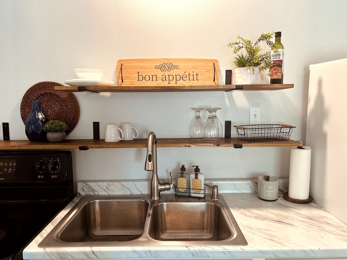 The kitchen area features a stainless steel sink beneath a marble countertop. Open wooden shelves display a 'bon appétit' sign, decorative items, glassware, and a wire basket. A bottle of oil and a candle are also visible, adding utility and charm.