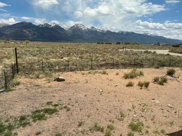 An open landscape features a gentle slope leading to a fence that borders the expansive property. The background showcases mountain peaks covered with snow under a partly cloudy sky, while sparse vegetation, including grass and low shrubs, covers the ground.
