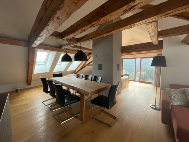 A spacious dining area features a long wooden table surrounded by black chairs. Exposed wooden beams add character to the ceiling. Natural light floods through the large windows, offering views of the surrounding landscape. A comfortable seating area is seen in the background.