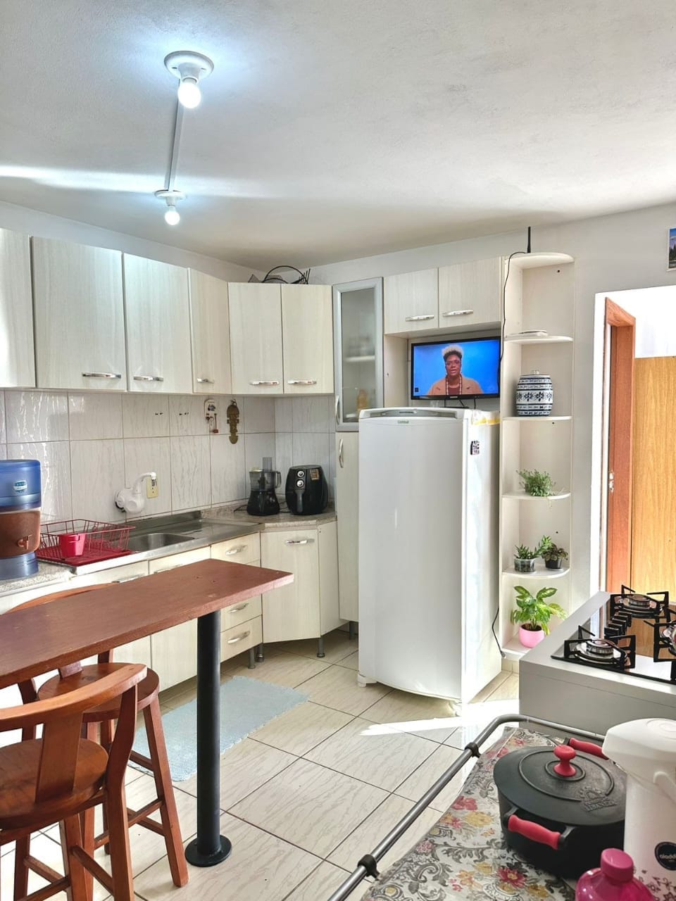 The kitchen features white cabinetry with a compact bar area and two wooden stools. A refrigerator and a television are visible, with a water dispenser and various kitchen appliances arranged neatly. A small plant adds a touch of greenery to the space.