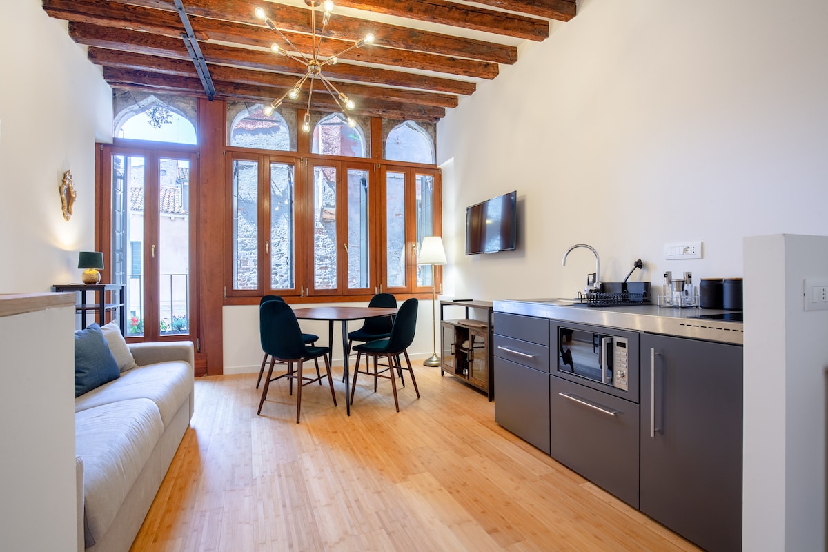 The living area features tall windows with wooden frames allowing natural light to flood the space. A round dining table is surrounded by four chairs. A modern kitchen with sleek cabinetry and appliances is visible, complemented by a light-colored sofa positioned nearby.