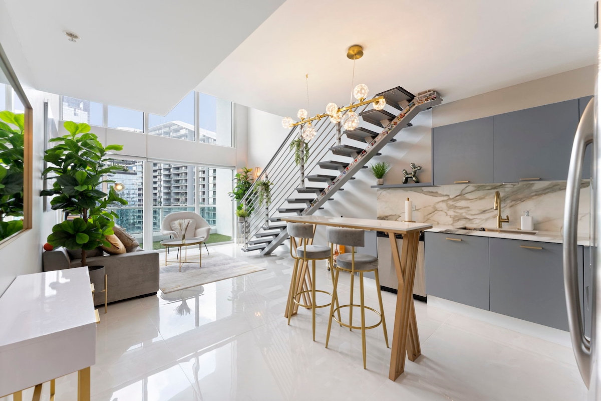 A modern loft space showcases a spiral staircase leading to an upper level. Natural light fills the room through expansive windows, with a small dining area featuring tall wooden stools and a sleek table. Green plants add a touch of vibrancy, complemented by stylish furnishings.