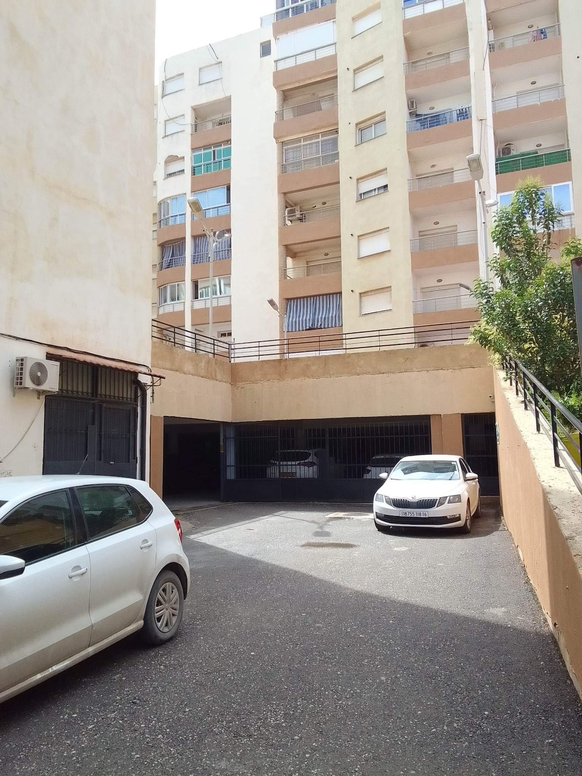 A secured parking area is shown with a smooth asphalt surface, featuring several designated parking spaces. A white vehicle is visible, parked near the entrance of the building, which has multiple stories. Surrounding walls and greenery create a secluded, private environment.