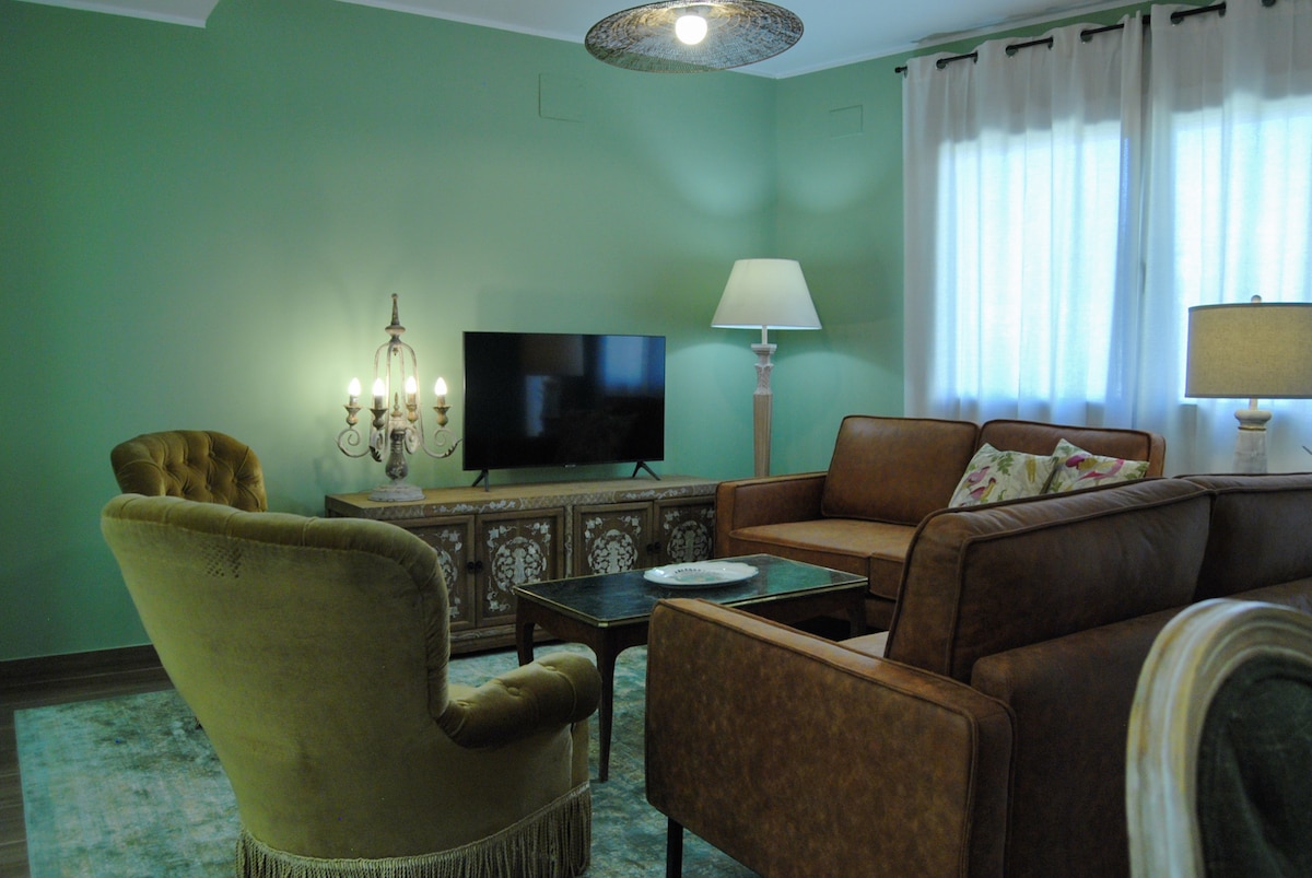 An inviting living area features a blend of seating options, including a velvet armchair and leather sofas. A coffee table occupies the center, with a decorative piece resting atop it. Soft lighting is provided by a lamp and wall sconces, creating a calm ambiance.