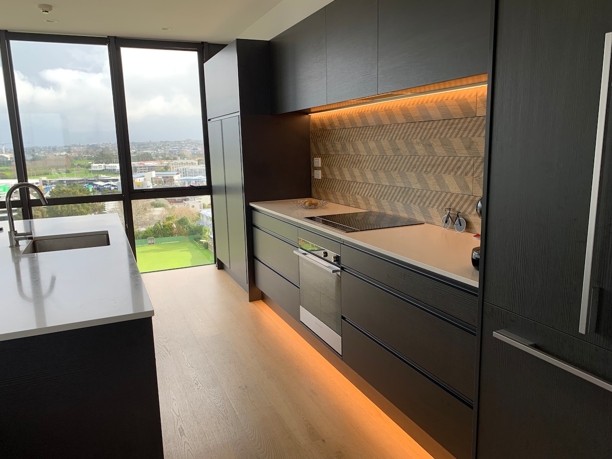 A modern kitchen features dark cabinetry and a chic herringbone backsplash. Under-cabinet lighting provides a warm glow along the countertop, which is equipped with an oven and cooking surface. Large windows offer views of the surrounding area, enhancing the sense of space and light.