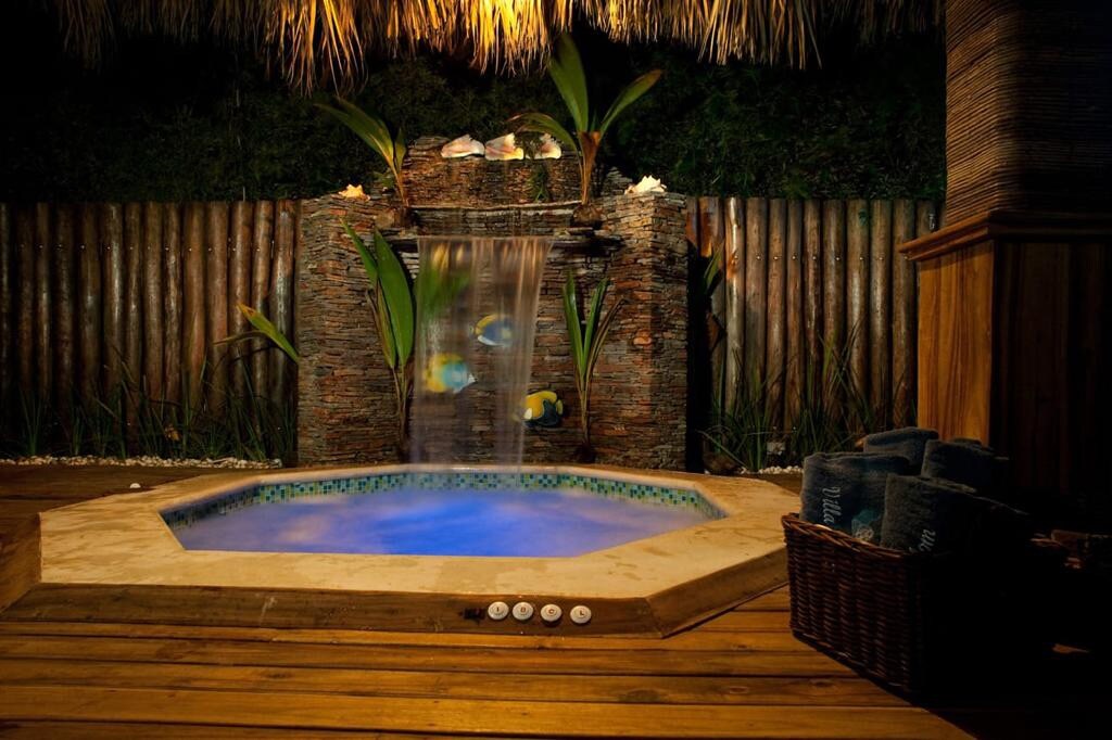 A relaxing hot tub is framed by a stone waterfall, surrounded by lush greenery. Soft blue lighting emanates from the water, enhancing the tranquil atmosphere. A woven basket containing towels is positioned nearby on the wooden deck, ensuring convenience for guests.