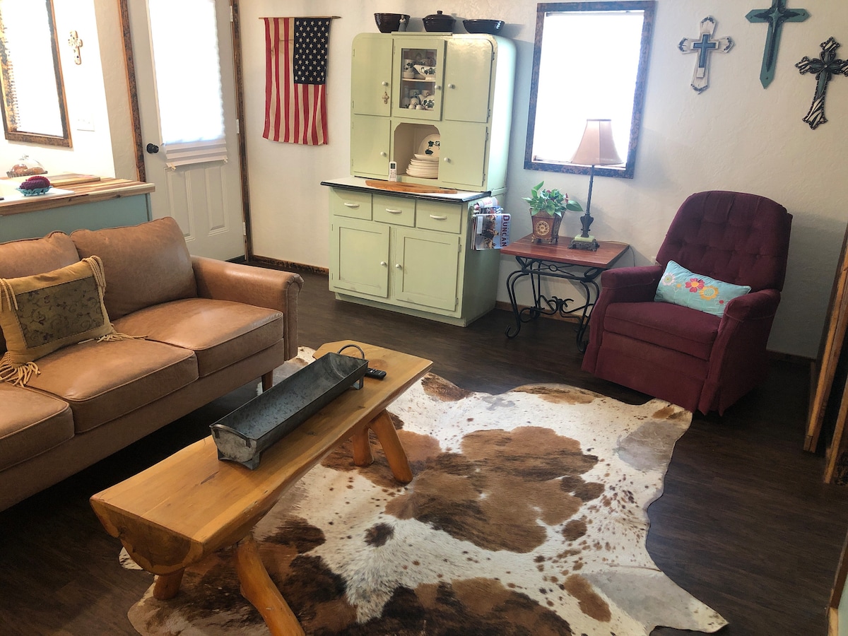 A living area features a brown couch paired with a wooden coffee table and a cowhide rug beneath. An accent chair in plum sits beside an old-fashioned sideboard, which is complemented by decorative items and framed mirrors on the walls. An American flag adds a touch of character.