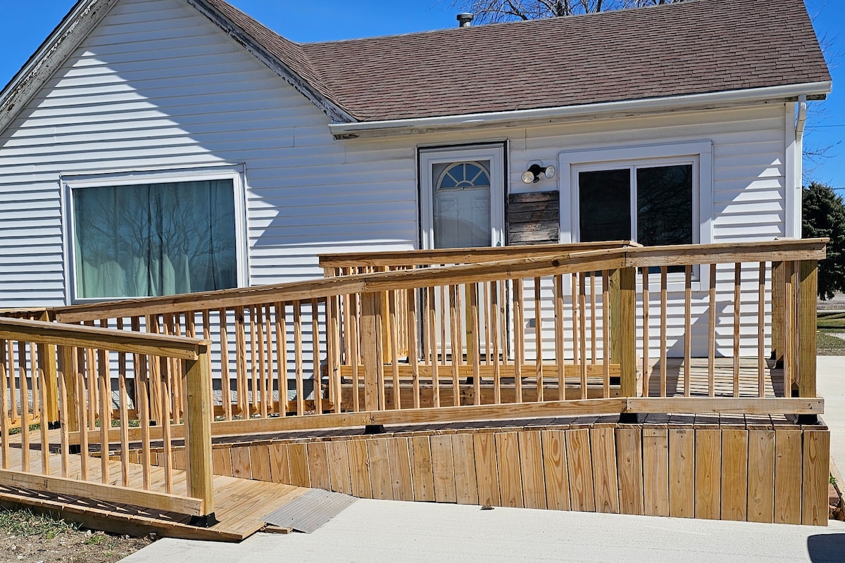 A wooden ramp leads up to the entrance of the house, providing accessibility. The front door is framed by a large window, allowing natural light to enter the space. The siding is white, and a clear blue sky is visible in the background.