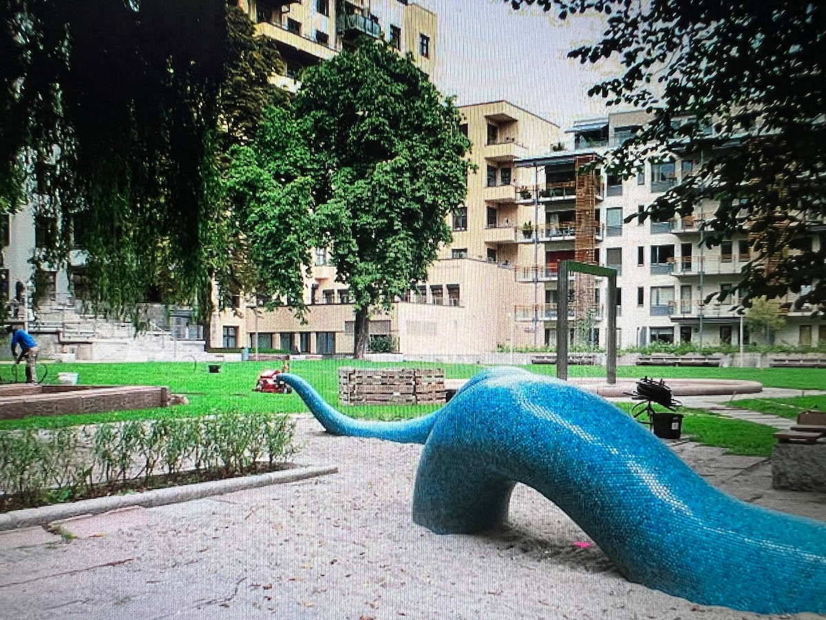A vibrant blue sculpture resembling a serpent is positioned on a sandy area within a landscaped park. Surrounding greenery includes large trees and manicured grass. In the background, modern apartment buildings are visible, integrating the urban setting with the natural elements.