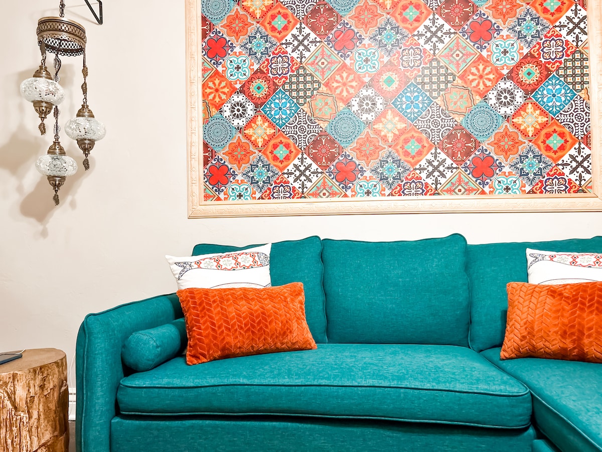 A vibrant turquoise couch is positioned against a colorful patterned wall art featuring intricate designs. Accent pillows in shades of orange add a pop of color. A natural wood side table enhances the cozy ambiance. Soft lighting is provided by decorative hanging fixtures.