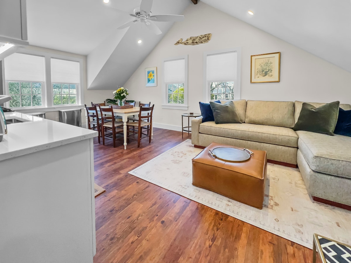 A bright living area features a spacious, light-brown sectional sofa alongside a coffee table. A dining table for four is situated by the windows, offering views of the outdoors. Light wooden floors create a warm ambiance, complemented by soft, neutral-toned furnishings.