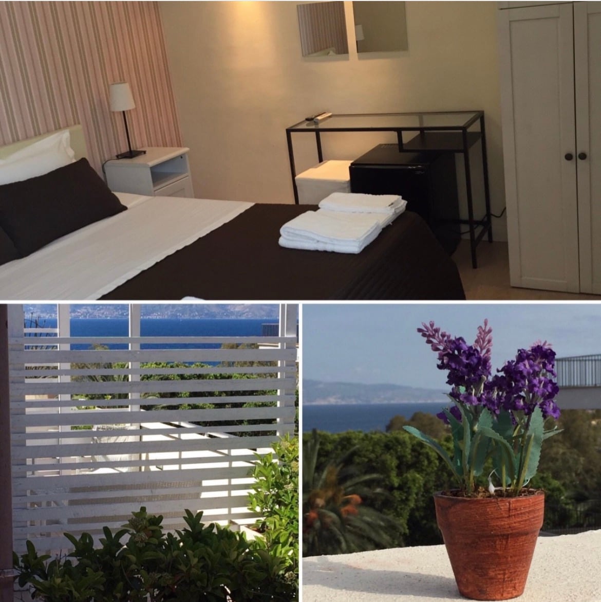 A well-appointed room features a spacious bed with neutral bedding and a bedside lamp. A small desk and chair are positioned nearby. In a separate view, a potted purple flower is placed on a terrace railing, next to greenery, overlooking scenic views of the sea.