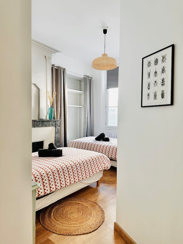 Cette chambre offre deux lits séparés. Les murs blancs et les rideaux gris créent une atmosphère douce et apaisante. La décoration simple, avec une cheminée et des touches naturelles, ajoute du charme à l'ensemble.