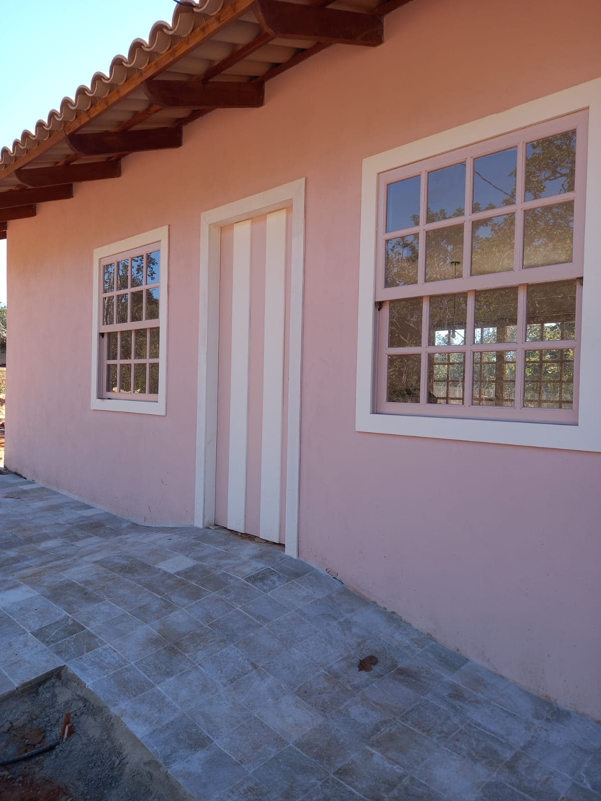 The exterior of the house is depicted, featuring a soft pink façade with two multi-paned windows on either side of a central white door. A paved pathway leads to the entrance, framed by natural surroundings visible through the windows.