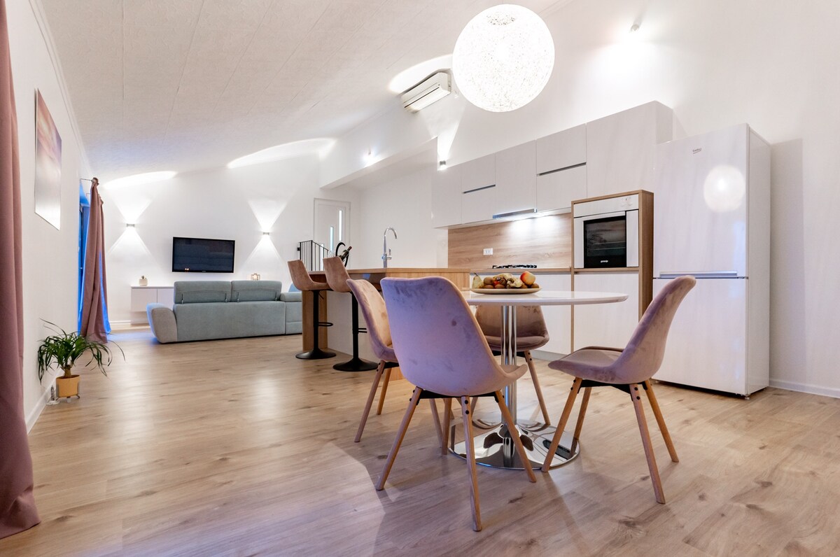 A modern dining area features a round table surrounded by four upholstered chairs. Light wood flooring extends throughout the space, complemented by a large white refrigerator and a sleek kitchen installation. A comfortable seating area is visible in the background, facing a wall-mounted television.