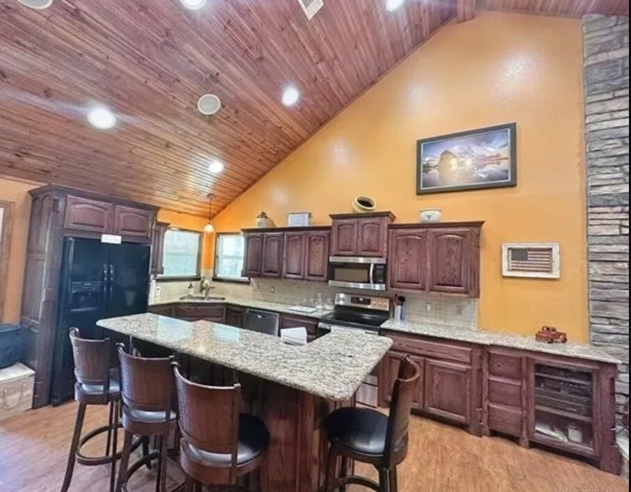 The kitchen features a high wood ceiling with recessed lighting, highlighting the granite countertops and dark wood cabinetry. A central island accommodates four barstools, while stainless steel appliances, including an oven and refrigerator, complement the warm color scheme.