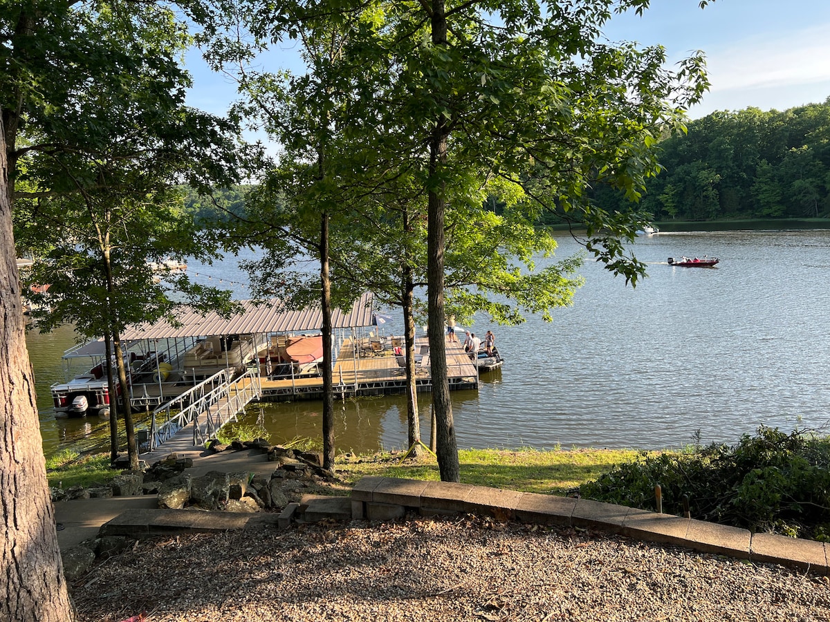A serene lakeside scene is captured, showcasing a shared boat dock with a slide, surrounded by lush trees. Several people can be seen enjoying the water. The calm lake reflects the greenery, creating a peaceful environment.