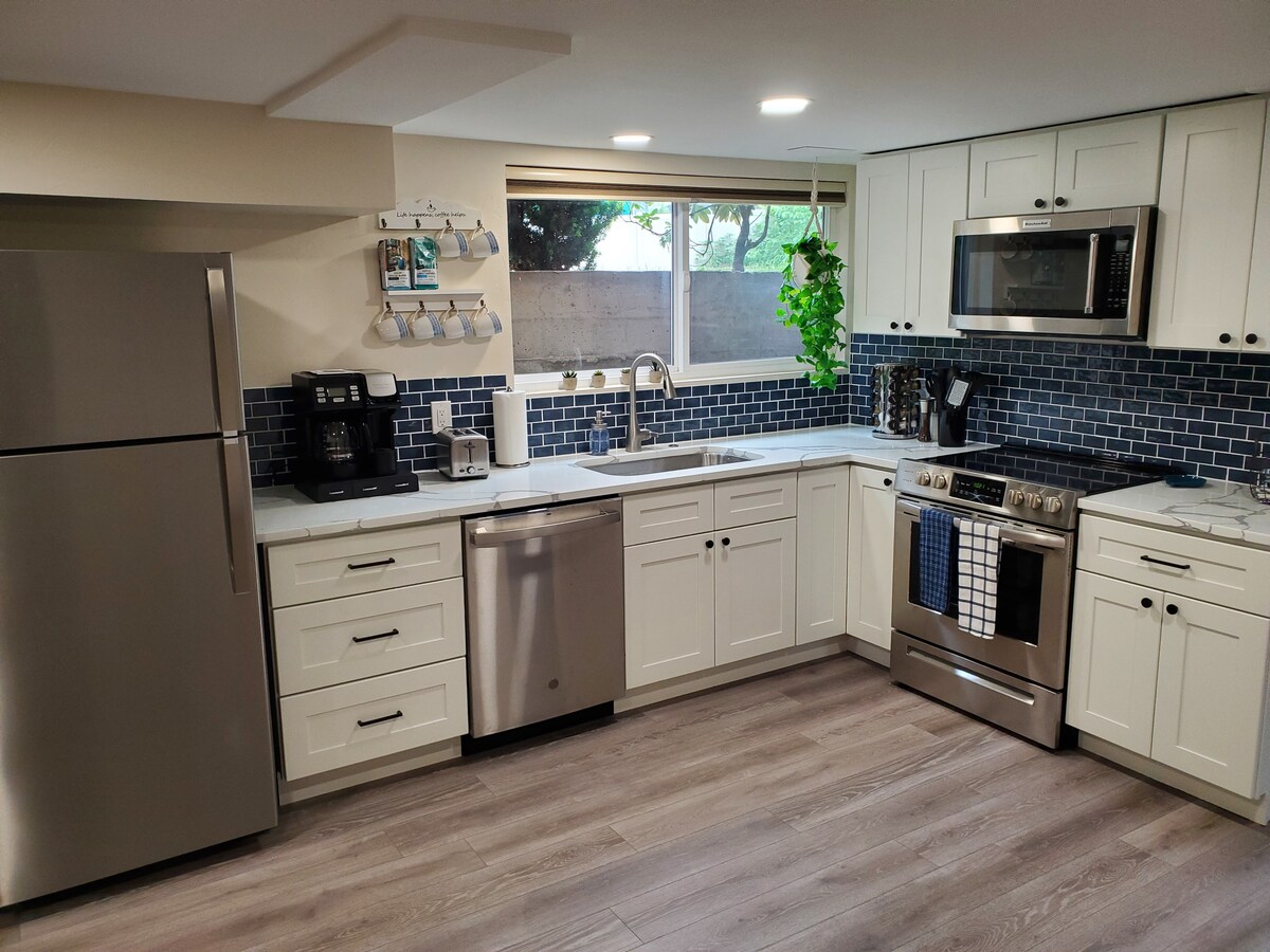 The kitchen features modern white cabinetry paired with a sleek stainless steel refrigerator and appliances. A blue tile backsplash adds a pop of color. Natural light enters through a window above the sink, complemented by plants arranged along the countertop.
