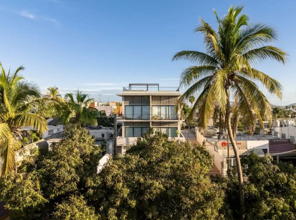 The image showcases a modern multi-story building surrounded by lush green trees, with palm trees prominently featured in the foreground. The clear blue sky provides a serene backdrop to the contemporary architecture, highlighting the structure's balconies and floor-to-ceiling windows.