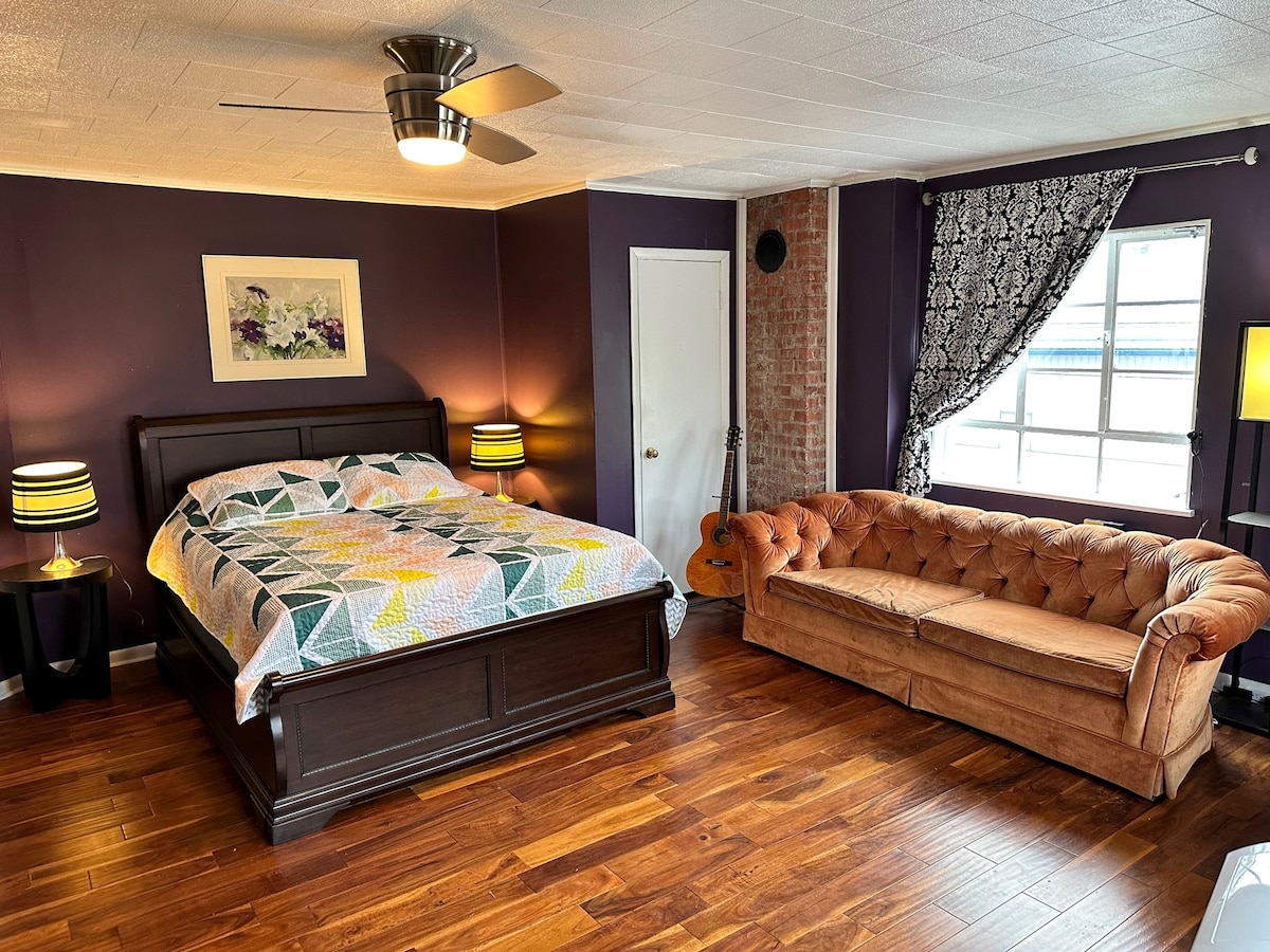 A spacious sleeping area features a dark wooden bed topped with a patterned quilt. Two bedside lamps provide soft lighting, with a modern ceiling fan overhead. A tufted brown couch is positioned nearby, complemented by a decorative curtain and large window allowing natural light.
