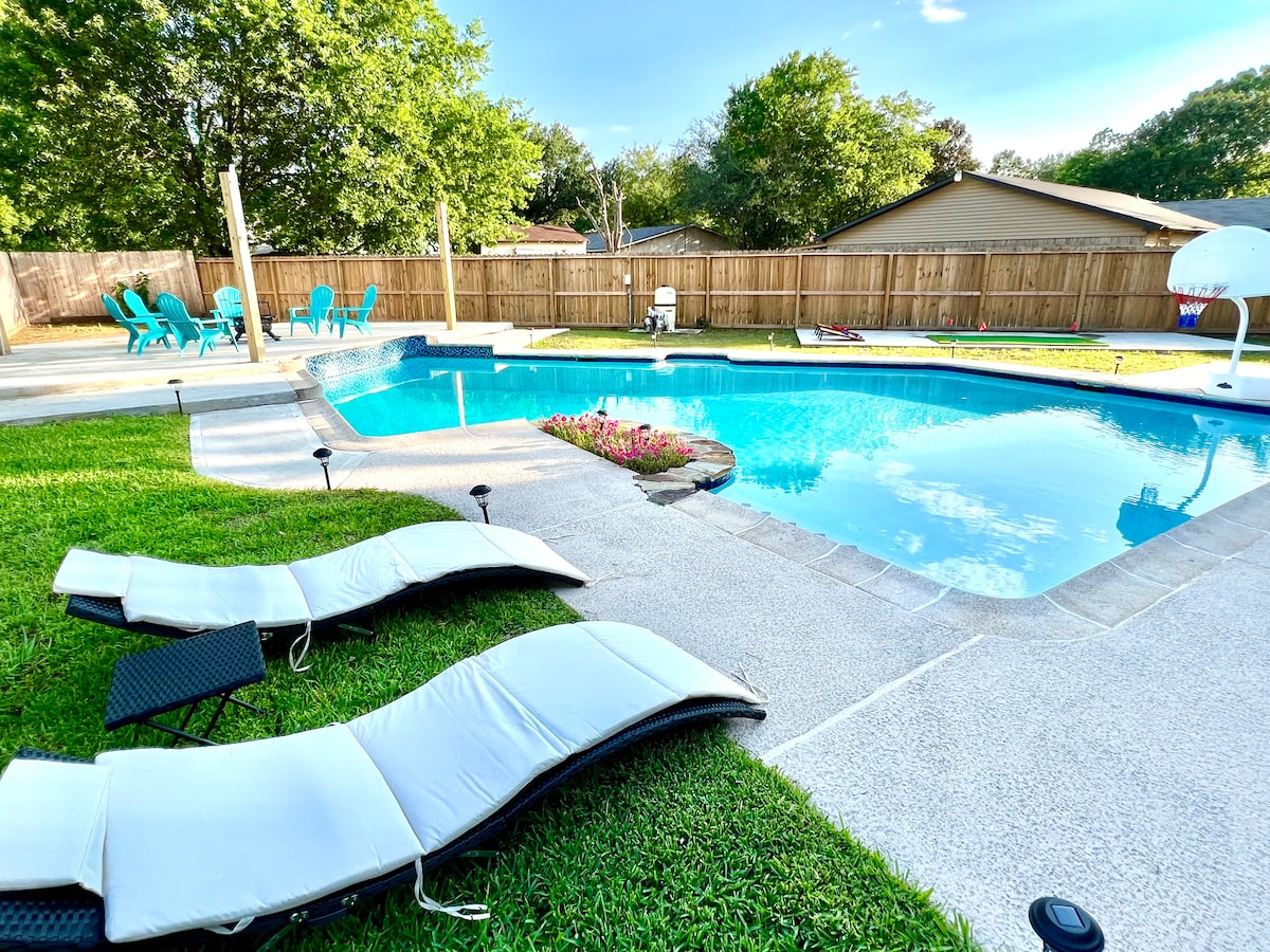 The outdoor area features a sparkling pool surrounded by a well-maintained yard. Lounge chairs are positioned close to the water, and colorful seating options are visible in the background. Green trees border the space, providing a sense of privacy.