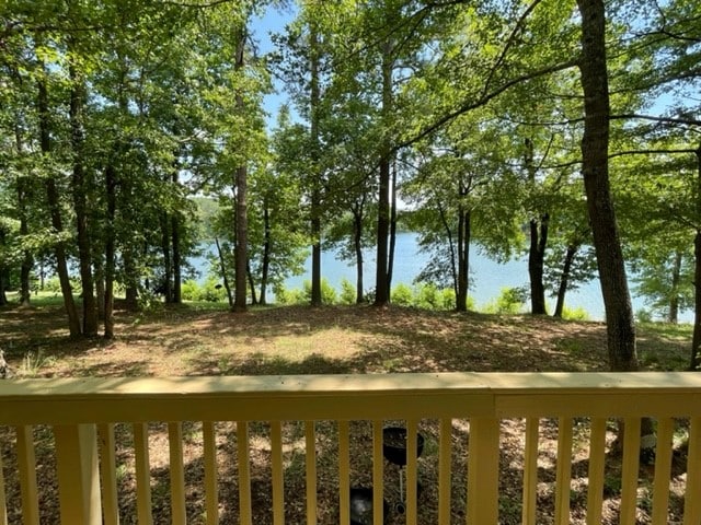 A serene view from the cabin's balcony showcases a picturesque lakeside landscape. Tall trees frame the scene, with sunlight filtering through the leaves. The calm water is visible in the background, creating a peaceful atmosphere suitable for relaxation.