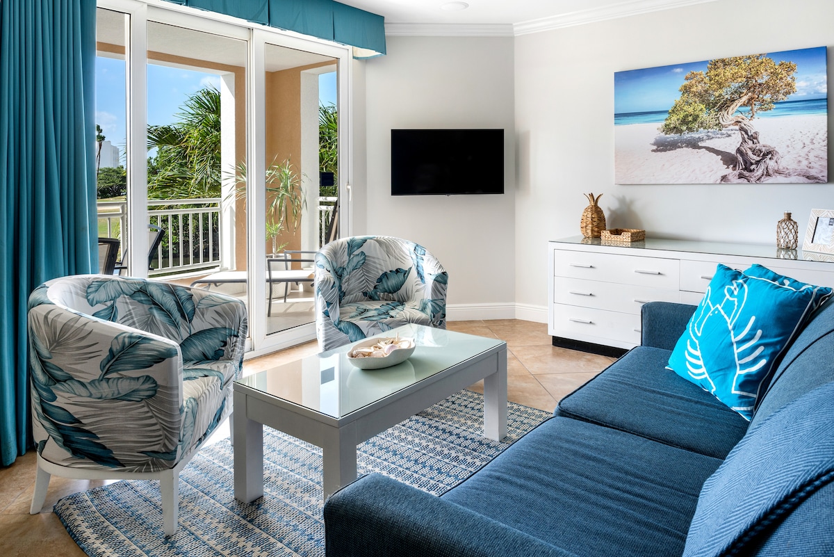 A light-filled living area showcases a comfortable dark blue sofa and two patterned chairs with tropical foliage designs. A modern coffee table occupies the center, while a large artwork depicting a beach scene is displayed on the wall. Sunlight streams in through sliding glass doors, leading to a private balcony.