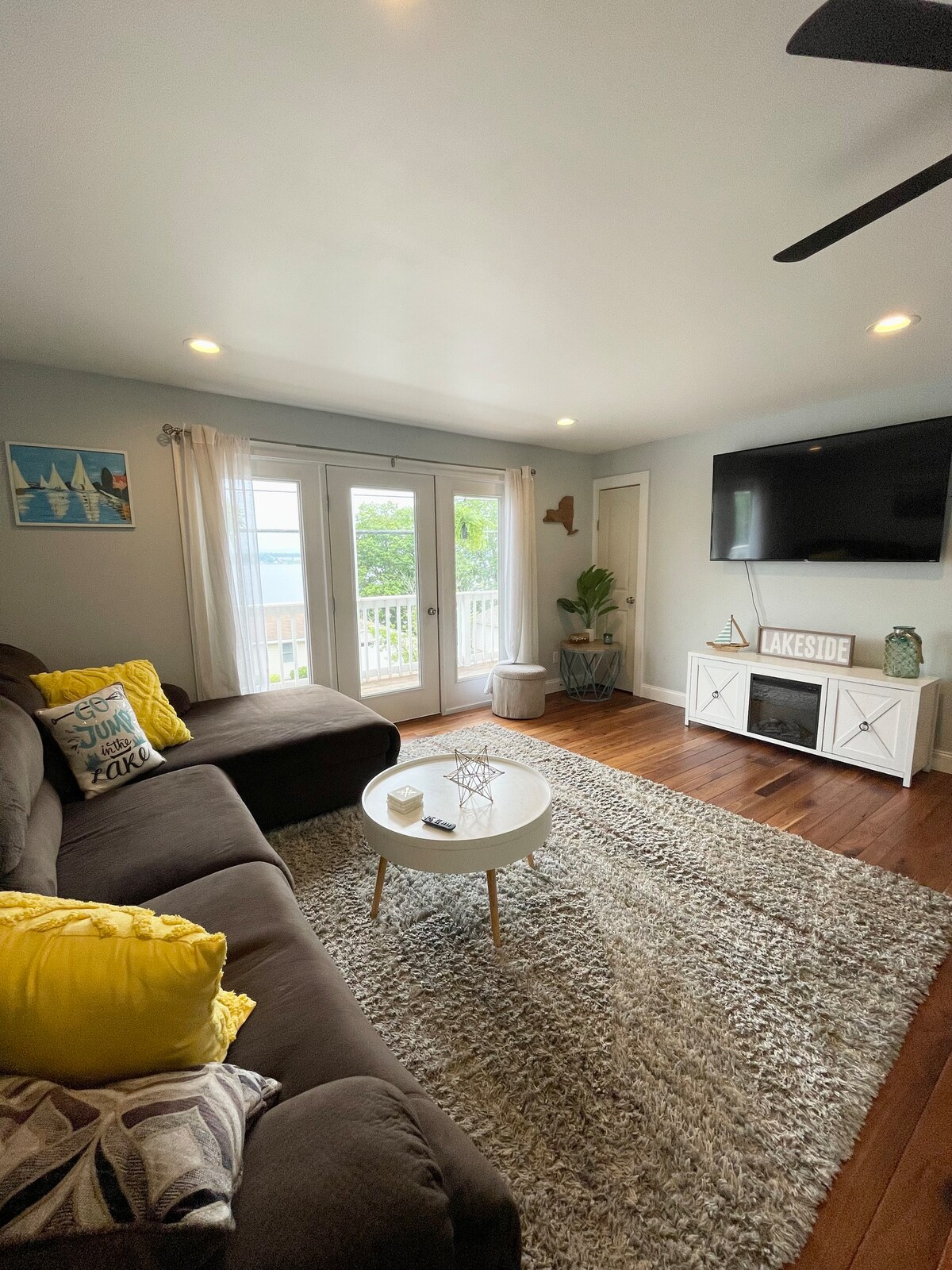 A cozy living area features a dark sectional couch adorned with yellow accent pillows. A light area rug covers the wooden floor, and a coffee table sits in front. Large glass doors allow natural light and scenic lake views, complemented by a mounted flat-screen TV.