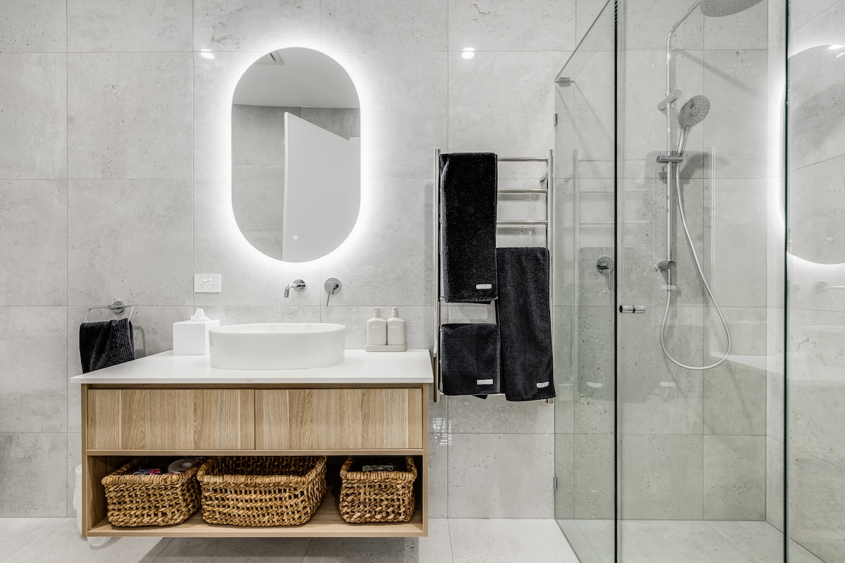 A modern bathroom features a sleek, circular mirror with backlighting above a minimalist vanity. A spacious shower enclosed in glass is visible, complemented by contemporary fixtures. Warm wooden accents are provided by a cabinetry unit with woven baskets, while plush black towels hang neatly on a rack.