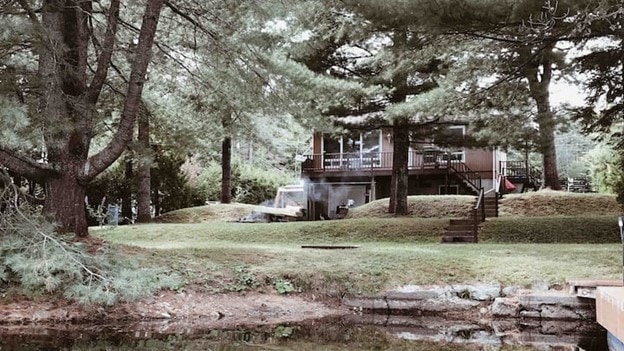 A wooden chalet is nestled among tall trees, overlooking a calm lake. A spacious lawn is visible, leading to a set of stairs that provide access to the entrance. The deck features a partially covered sitting area, enhancing the connection with nature.