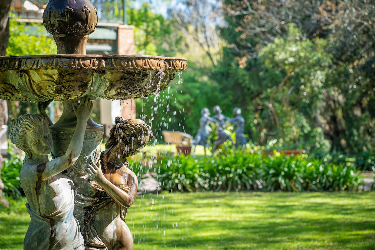 A decorative fountain featuring intricately carved figures stands amidst a lush garden. Water gently cascades from the fountain, creating a soothing ambiance. Green foliage surrounds the area, with sculptures visible in the background, providing a serene outdoor setting.