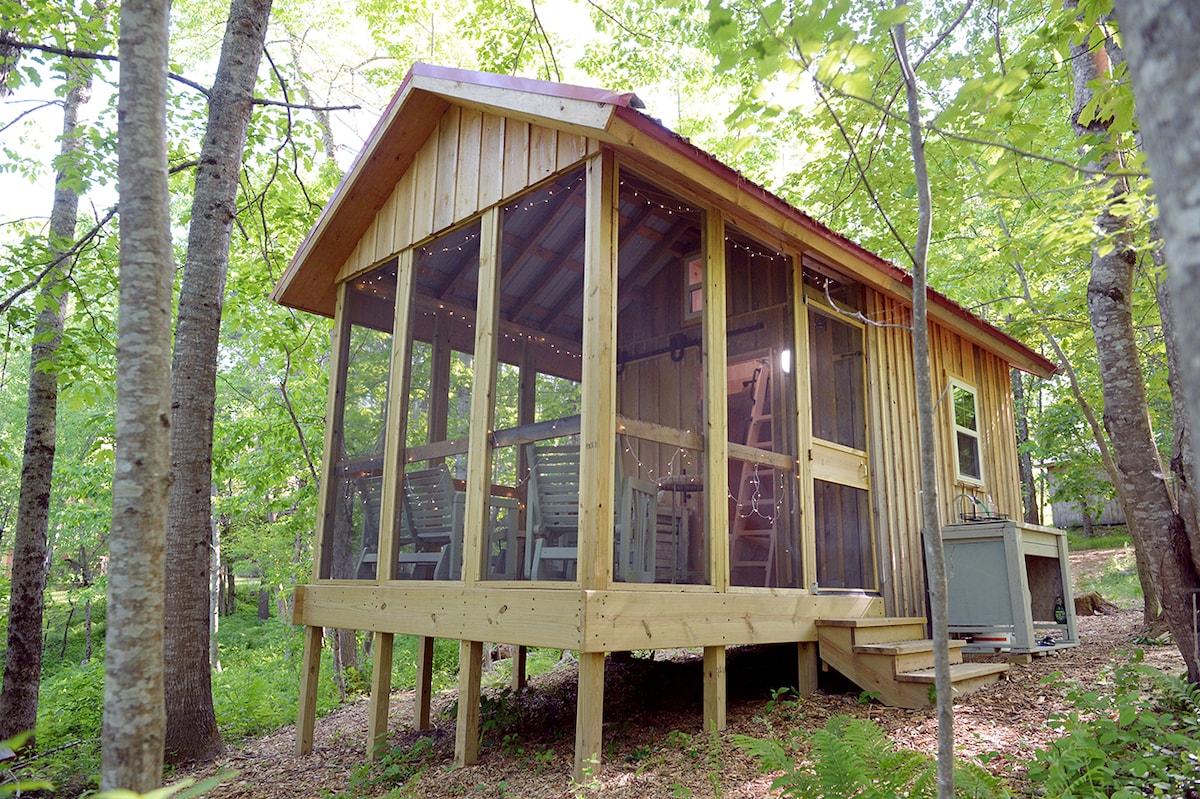 The cabin, elevated on wooden stilts, is surrounded by lush greenery. Screened windows allow natural light to filter in, illuminating the inviting interior spaces. Twinkling lights adorn the screened porch, creating a cozy atmosphere as the cabin blends harmoniously with its woodland setting.