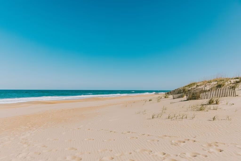 A sandy beach stretches towards the horizon under a clear blue sky. Gentle waves lap against the shore, while a line of grassy dunes is visible in the distance, bordered by wooden fencing. Footprints are imprinted in the soft sand, suggesting recent visitors.