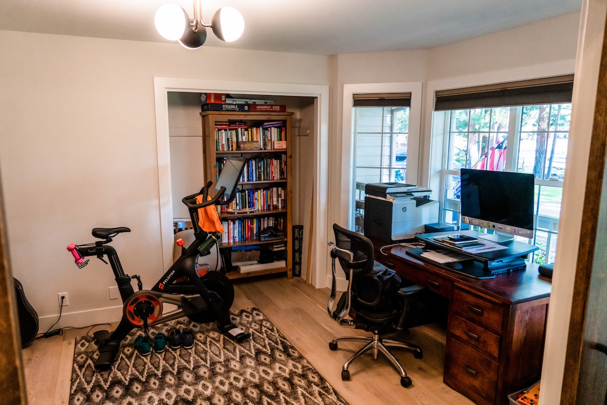 Канцеларија со биро за стоење, велосипед Peloton, тегови, подлоги за јога и удобен брачен кревет