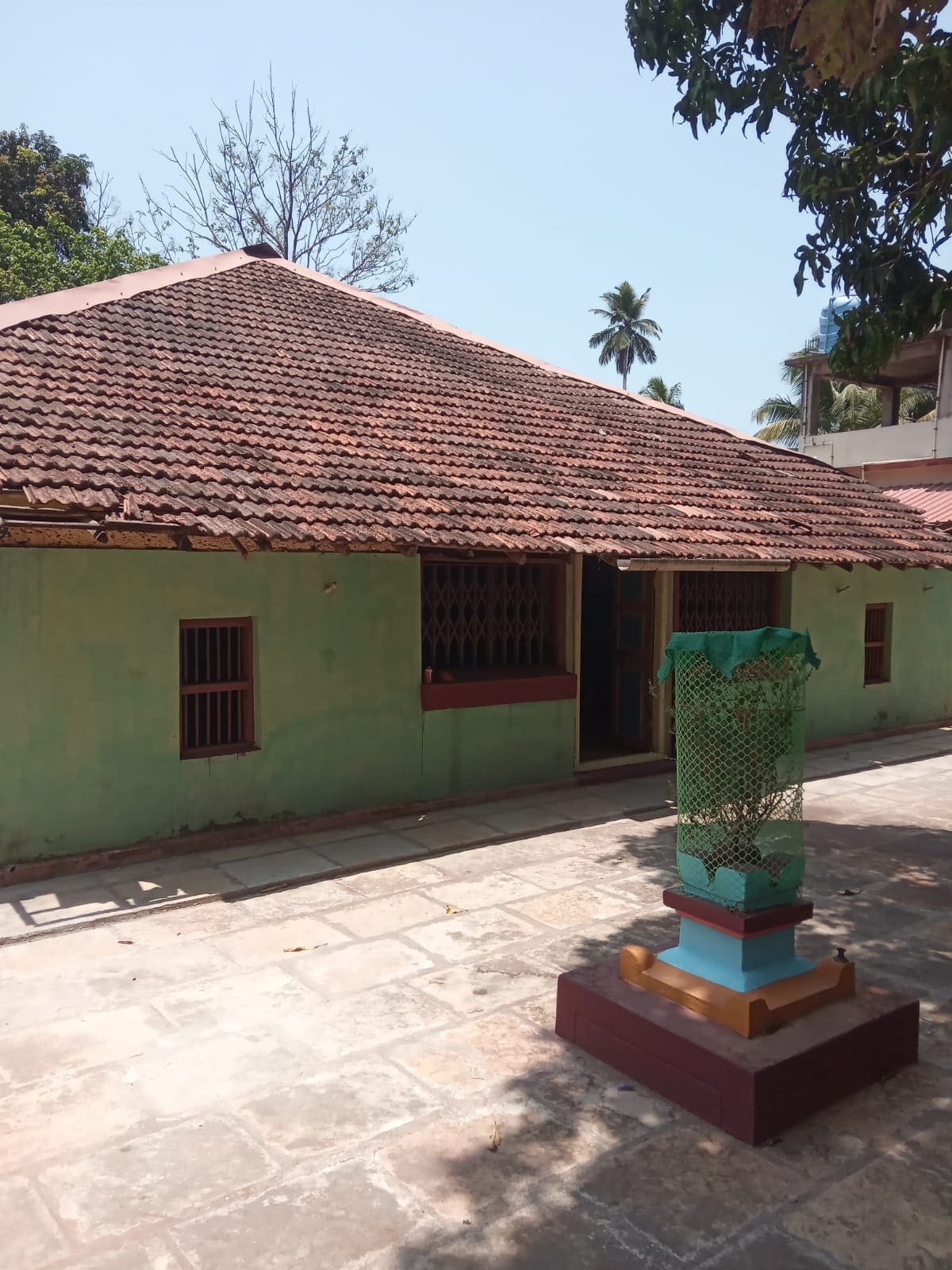 A rustic mud house with a sloped terracotta roof is presented in a quaint outdoor setting. The exterior walls are painted in a soft green hue, adorned with wooden window grilles. In the foreground, a decorative lantern stands on a raised pedestal, surrounded by a tiled courtyard.