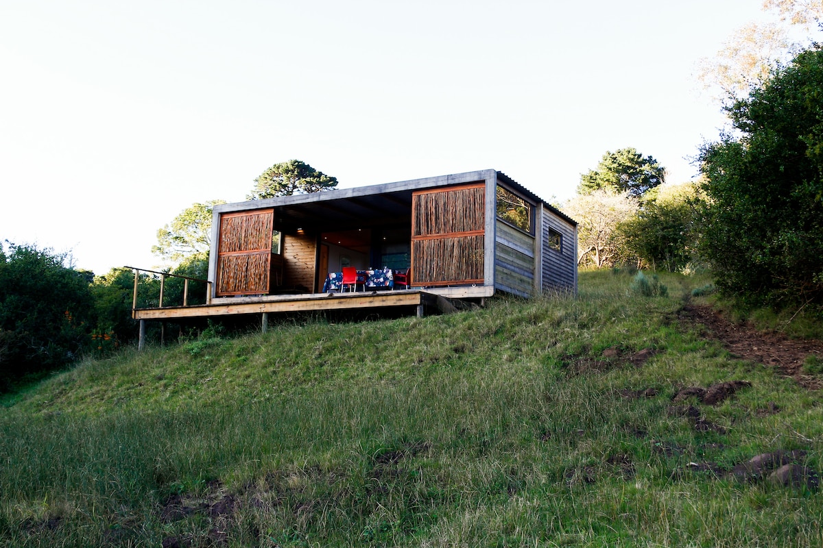 The cabin is nestled on a hillside surrounded by lush greenery. Large glass doors open to a wooden deck, where a dining table and chairs offer an outdoor eating space. The structure features a modern design with natural wood accents, blending harmoniously with the landscape.