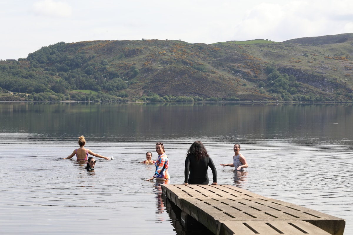 A calm lake scene features a wooden dock extending into the water, with individuals enjoying a swim. Lush green hills rise in the background, creating a serene atmosphere. The clear water reflects the gentle colors of the sky, inviting relaxation and connection with nature.
