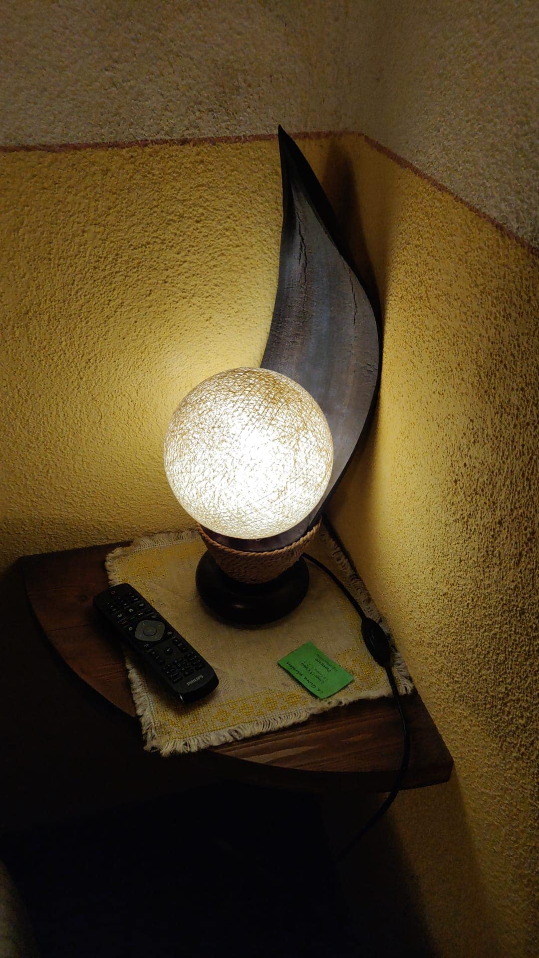 A unique lamp with a textured globe illuminates a small bedside table. A decorative wooden piece is positioned next to the lamp, while a remote control and a small green note are placed on the table, creating a functional yet distinct corner.