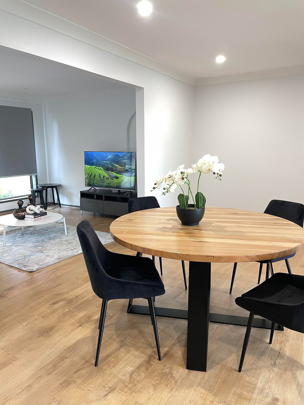 A circular wooden dining table is set in a modern dining area, surrounded by four dark chairs. A potted orchid decorates the table's center, while a television and light-colored furniture are visible in the background, contributing to an open and inviting space.
