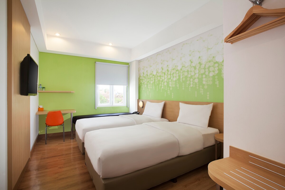 A bright sleeping area features two spacious beds with crisp white linens. A vibrant green accent wall adds color to the room. Natural light enters through a window, illuminating a small workspace with an orange chair and a wooden shelf.