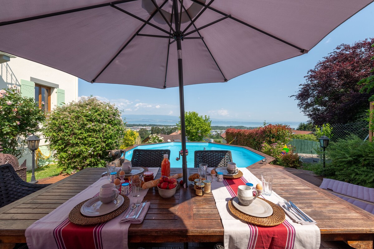 A spacious outdoor dining area is set beneath a large umbrella, featuring a wooden table adorned with tableware and fresh fruit. In the background, a pool reflects the clear sky, surrounded by lush greenery and flowers, creating a serene ambiance.