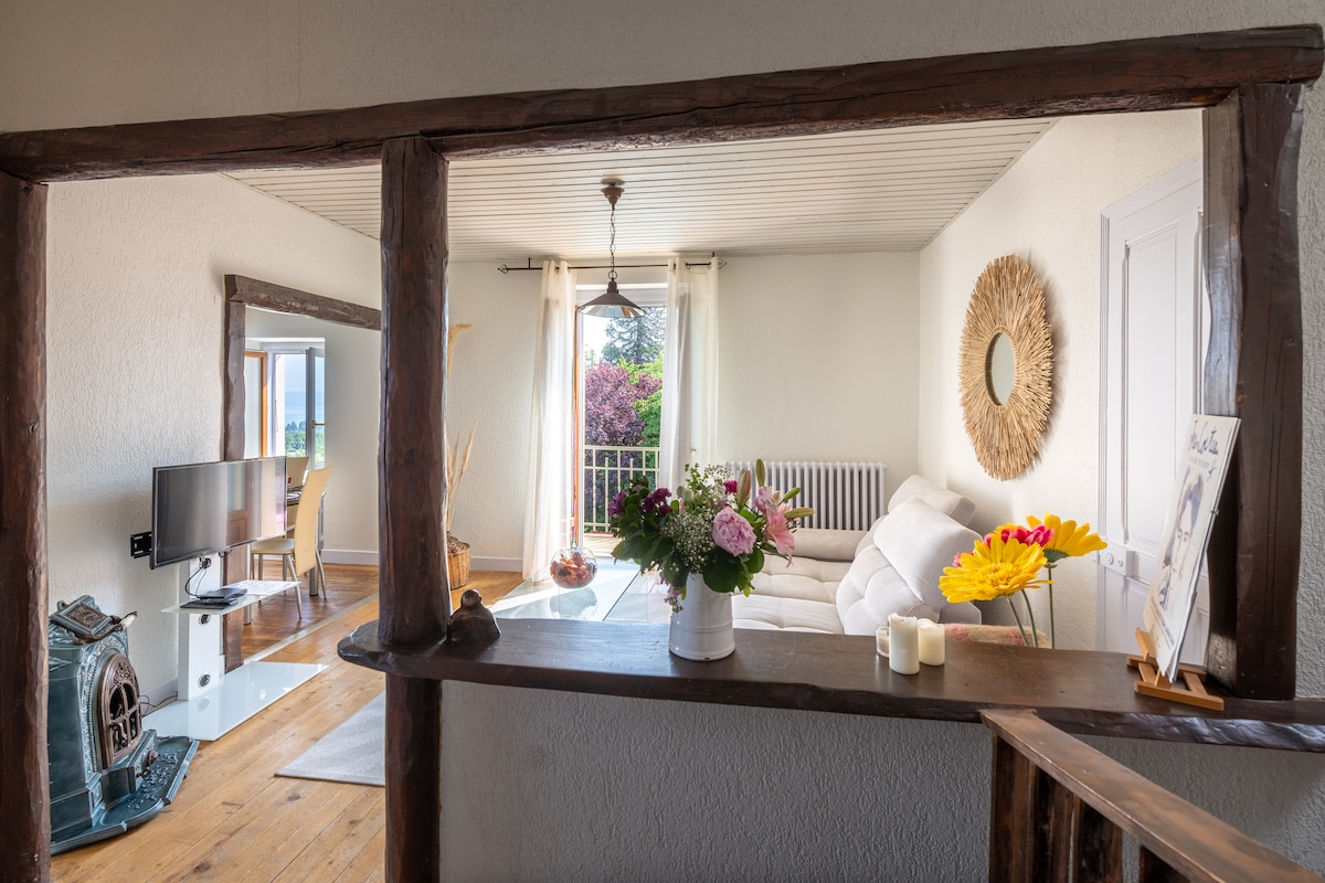 A view from an open doorway reveals a bright living area with neutral-toned walls and a comfortable white sofa. A television is positioned in the corner, while natural light streams through large windows, illuminating decorative elements such as flowers and a round mirror.
