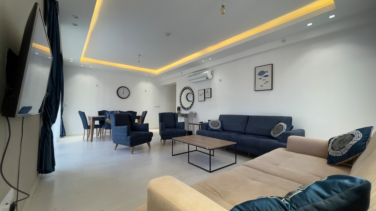 An open living area features a blend of modern furniture, including a comfortable sectional sofa and accent chairs. A central coffee table is placed over light-colored flooring. Bright wall art and ambient lighting highlight the inviting space, which is connected to a dining area with a large table.