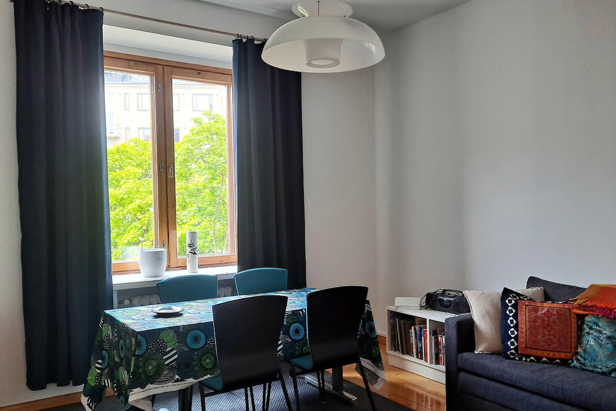 A dining area is highlighted by a table adorned with a colorful tablecloth. Four modern chairs surround it, while a large window invites natural light and offers views of greenery outside. A cozy couch adorned with decorative cushions is positioned against the wall.