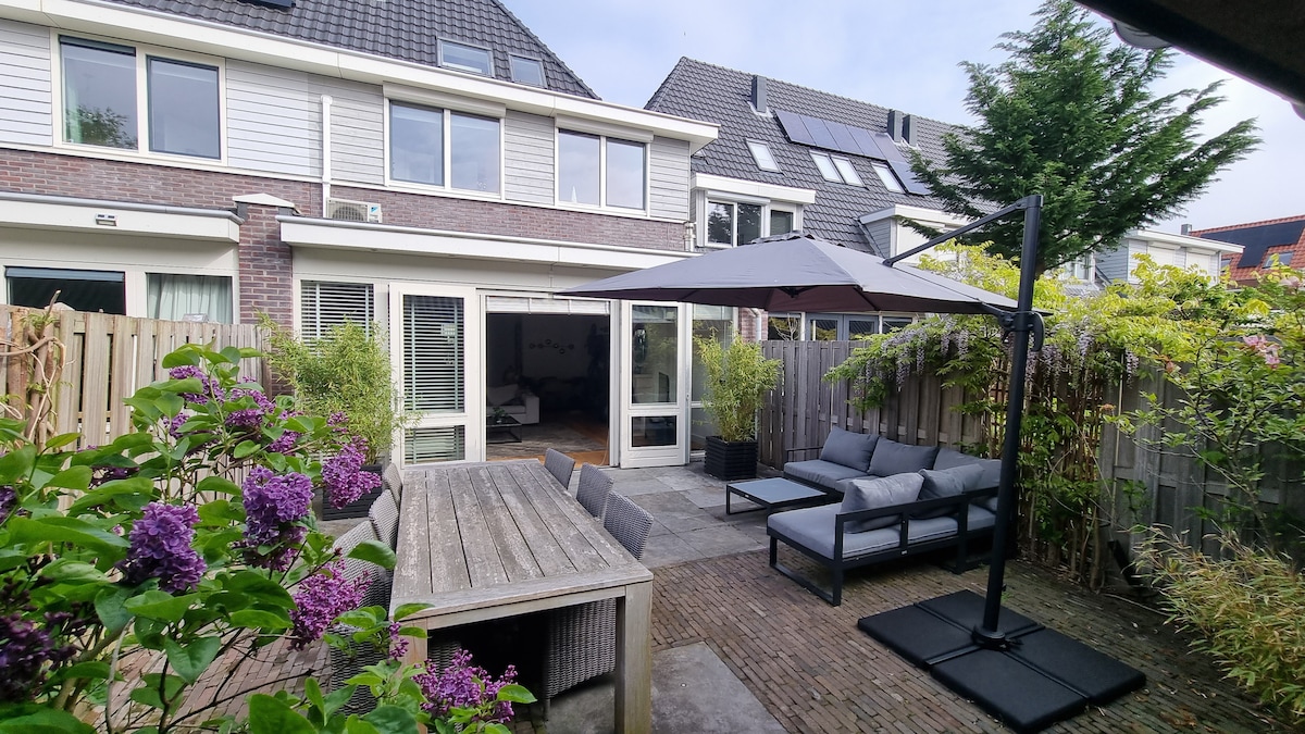 An outdoor area features a large dining table surrounded by plants and flowers. Comfortable seating arrangements are provided under a shaded umbrella. The private garden is accessed through glass doors, seamlessly connecting indoor and outdoor spaces.