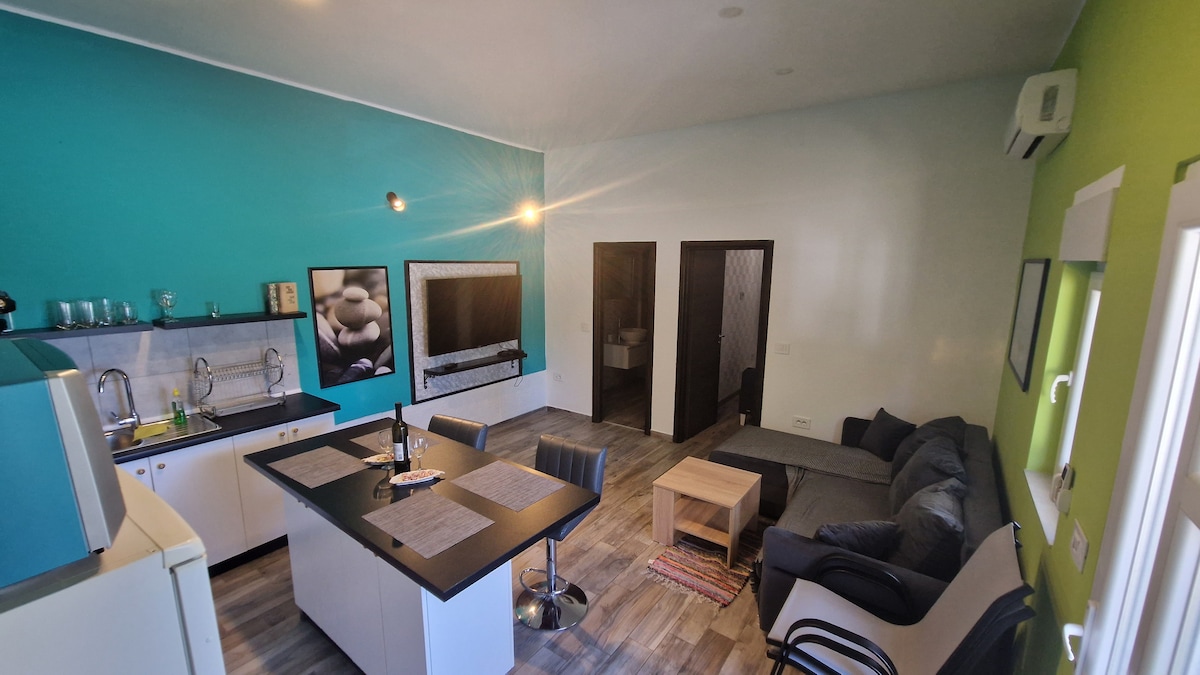 An open-plan living area is showcased, featuring a kitchen with modern appliances and a cozy seating area. The walls are painted in calm shades of turquoise and green. A dining table with chairs is positioned near the kitchen, creating an inviting atmosphere for meals and gatherings.